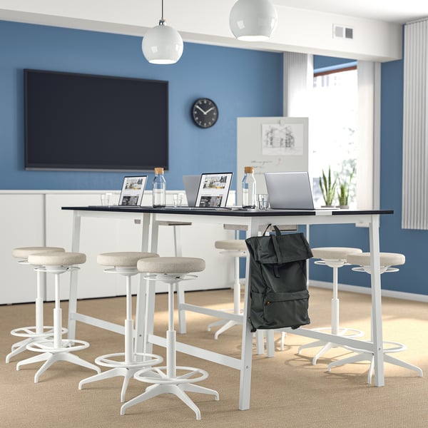 Moderna configuración de oficina con mesa de conferencias alta MITTZON y taburetes blancos. La mesa tiene computadoras portátiles, botellas y una mochila, con un televisor grande en la pared azul.
