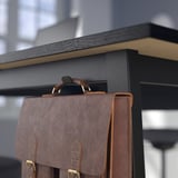 El bolso de cuero cuelga de un escritorio negro con reposapiés y módulo de gestión de cables.