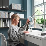 Un niño usa una tableta en un escritorio con un soporte que sostiene la tableta. El escritorio tiene una lámpara, una planta y otros artículos. La pared tiene estanterías con libros y cajas.