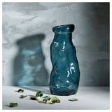 Jarrón de vidrio reciclado azul con diseño ondulado; Se asienta sobre una superficie blanca con hojas dispersas.