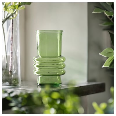 Jarrón de vidrio verde acanalado sobre una superficie reflectante, rodeado de plantas.