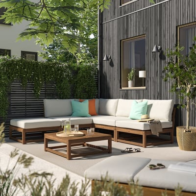 Patio moderno con sofá NÄMMARÖ, cojines de colores, mesa de madera, vegetación y edificios contemporáneos.