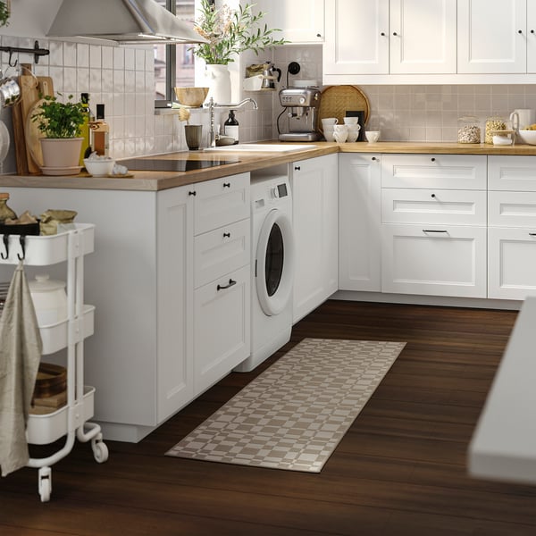 Cocina moderna con armarios blancos, encimeras de madera y alfombra NATTÅG. Cerca: lavadora, carrito, electrodomésticos.