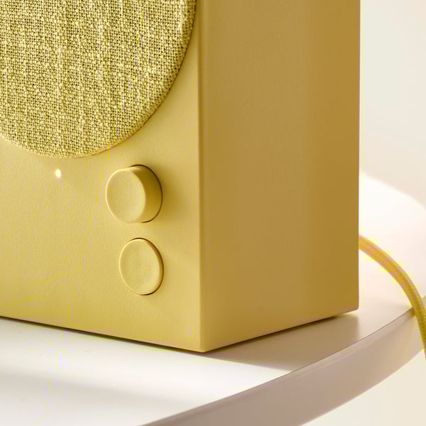 Primer plano de un elegante altavoz bluetooth verde oliva con cubierta de tela y controles redondos, colocado sobre una superficie curva y elegante.