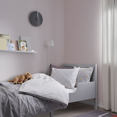 Una acogedora funda nórdica gris y blanca con estampado de margaritas NATTSLÄNDA, hecha de algodón, se adapta perfectamente a una cama pequeña en una habitación rosa. Encima hay un reloj de pared y libros.