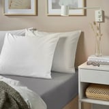 Cama con almohadas blancas de algodón Nonnea, sábanas grises, mesita de noche de madera, libros, jarrón y arte de pared.