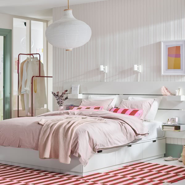 Dormitorio moderno con cama blanca NORDLI, almohadas rosas a rayas, lámpara colgante, alfombra a rayas rojas, perchero con prendas blancas y colorido arte mural.