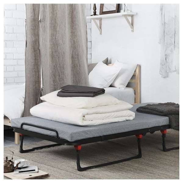 Dormitorio moderno con cama blanca, cama de invitados de tela gris, pies rojos, mantas dobladas encima. Cortinas blancas, estante de madera con espejo.
