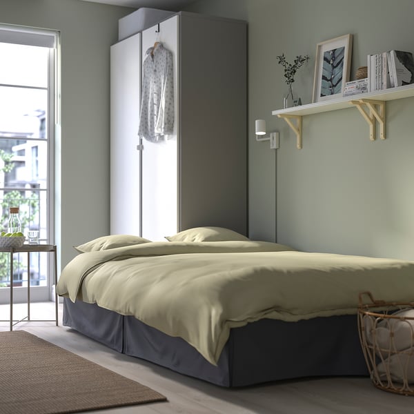 Acogedor dormitorio con cama beige, estanterías blancas, paredes verdes. La cama tiene falda gris, albornoz colgante, luz de pared y canasta al lado de la cama.