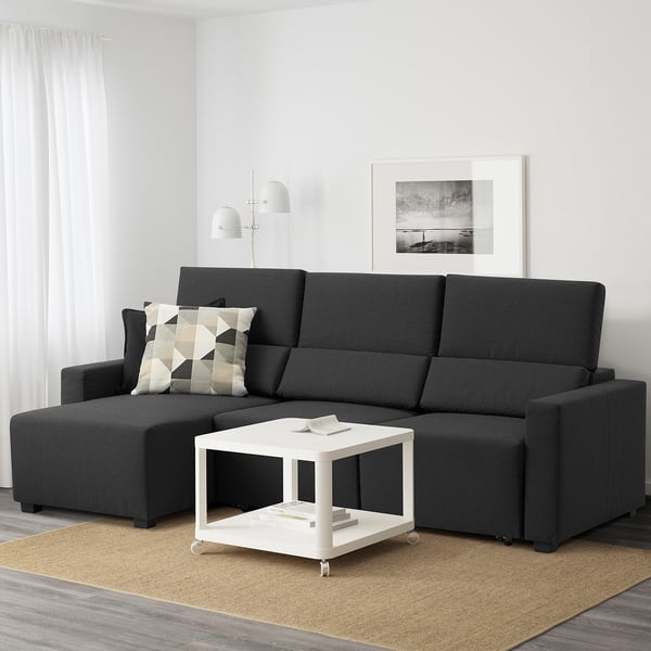 Sala de estar moderna con sofá negro en forma de L, tumbona ajustable, alfombra amarilla y mesa de centro con ruedas.