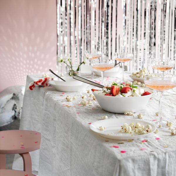 Mesa con cuencos y vasos OFTAST, vidrio templado blanco, palomitas de maíz esparcidas, fresas y confeti rosa, fondo plateado brillante.