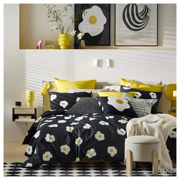 Dormitorio luminoso con ropa de cama blanca y negra, flores / almohadas amarillas, silla blanca, alfombra / mesa negra, jarrón, lámparas de pared, marcos.