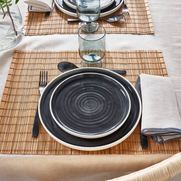 Mesa con platos blancos y negros, cubiertos oscuros, cristal, mantel individual tejido, servilleta beige.