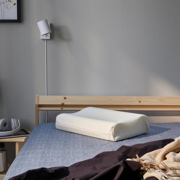 Un dormitorio minimalista, una almohada PAPEGOJBUSKE sobre una cama con cabecero de madera, una lámpara moderna y una pequeña mesita de noche con auriculares.