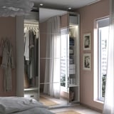 Dormitorio moderno con armario con espejo PAX. Es de color blanco crema, tiene puertas correderas y alberga ropa, con una ventana al lado.