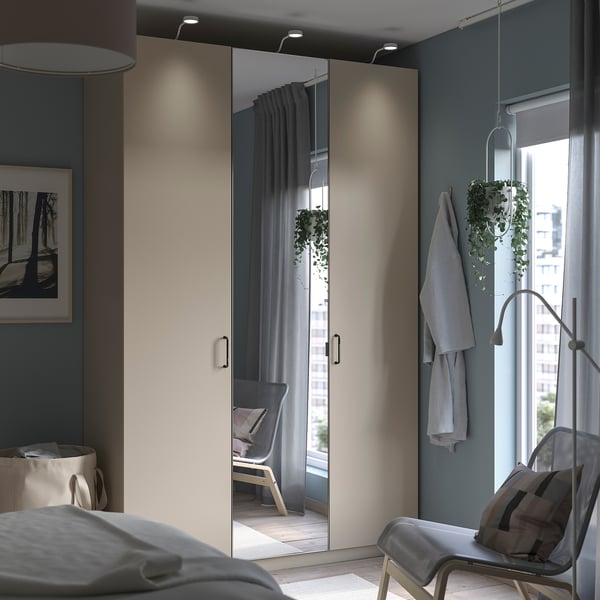 Un dormitorio minimalista, un armario PAX con puertas con espejos, una planta colgante y una acogedora silla junto a la ventana. La decoración gris acentúa el espacio.