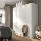 Dormitorio moderno con armario PAX blanco, tiradores dorados, cárdigan marrón, cama gris, silla de mimbre, armario alto.