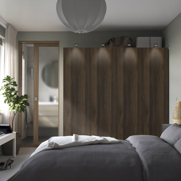 Dormitorio moderno, gran armario PAX de madera oscura con elegantes asas, bolsas en la parte superior, flanqueadas por cortinas. Una cama gris con sábanas blancas está en primer plano.
