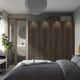 Dormitorio moderno, gran armario PAX de madera oscura con elegantes asas, bolsas en la parte superior, flanqueadas por cortinas. Una cama gris con sábanas blancas está en primer plano.