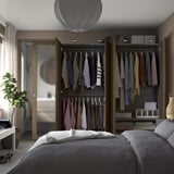 Armario con espejos con ropa, estantes, cajones, iluminación integrada y puertas de cierre suave. Ropa de cama gris sobre cama parcialmente visible.