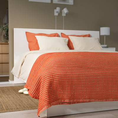 Cama doble con colcha de tejido gofre PELARTUJA naranja, almohadas blancas, cabecero y mesita de noche lámpara.