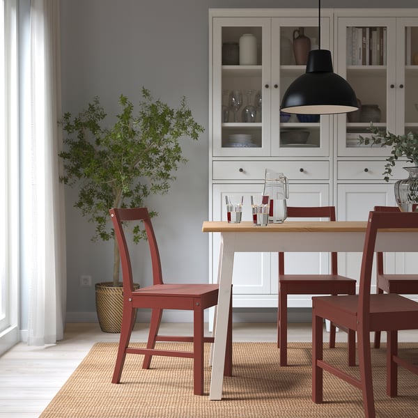 Silla PINNTORP de madera roja con respaldo inclinado, alrededor de la mesa de comedor sobre alfombra, mueble blanco con cristalería, planta. Acogedor, minimalista, rústico-moderno.