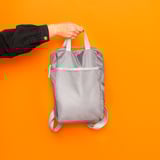 Persona sosteniendo una mochila de malla gris contra una pared de color naranja brillante, mostrando su diseño y correas ajustables.