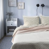Colcha acolchada blanca con almohadas, mesita de noche con libros y jarrón.
