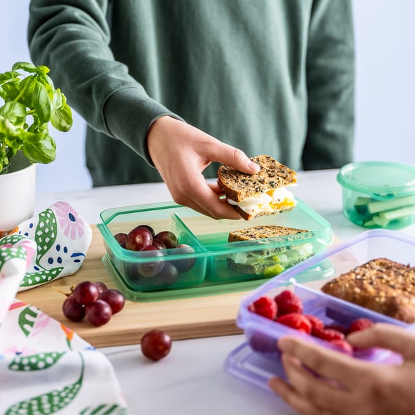 Persona que toma un sándwich de una lonchera compartimentada llena de fruta y un bocadillo en una tabla de madera con varios recipientes alrededor.