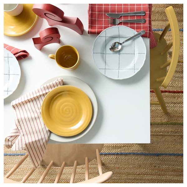 Platos y tazas de color amarillo brillante sobre manteles individuales a rayas, colocados sobre una mesa blanca con sillas rústicas.