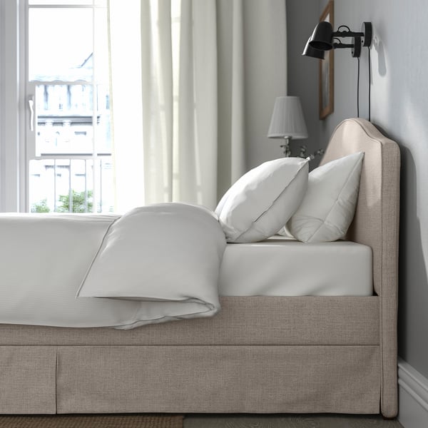 Cama con cabecero curvado, almohadas blancas y funda de color beige claro. Lámpara de pared y ventana con vista a la ciudad.
