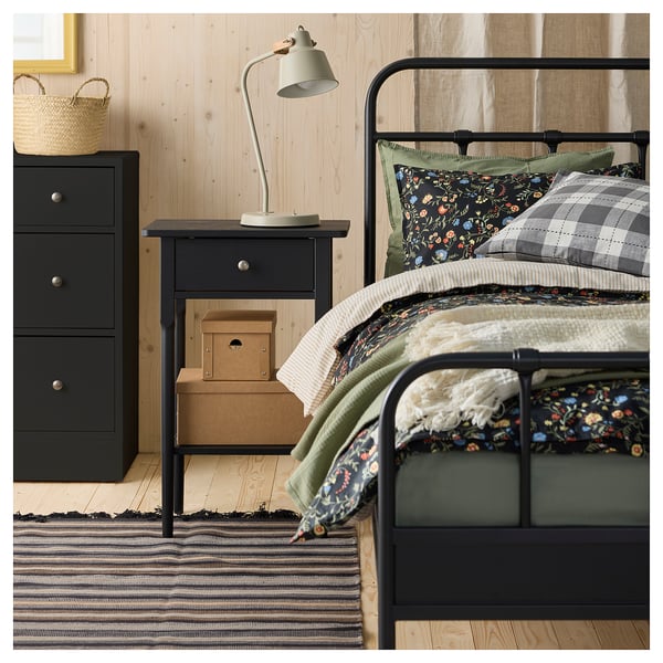 Dormitorio con cama de metal negro, ropa de cama floral y a cuadros, mesilla de noche de madera con lámpara y alfombra a rayas blancas y negras.