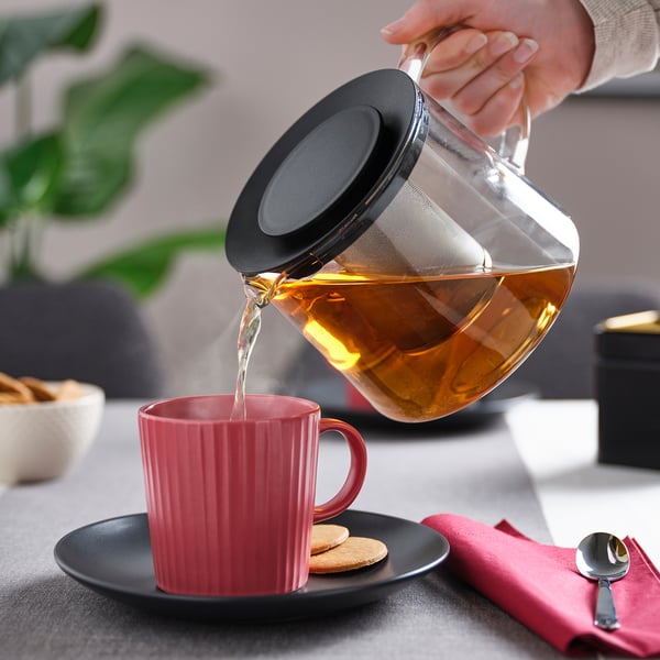 La tetera vierte el té en una taza roja en un plato oscuro, con infusor integrado, el vapor sale de la boquilla.