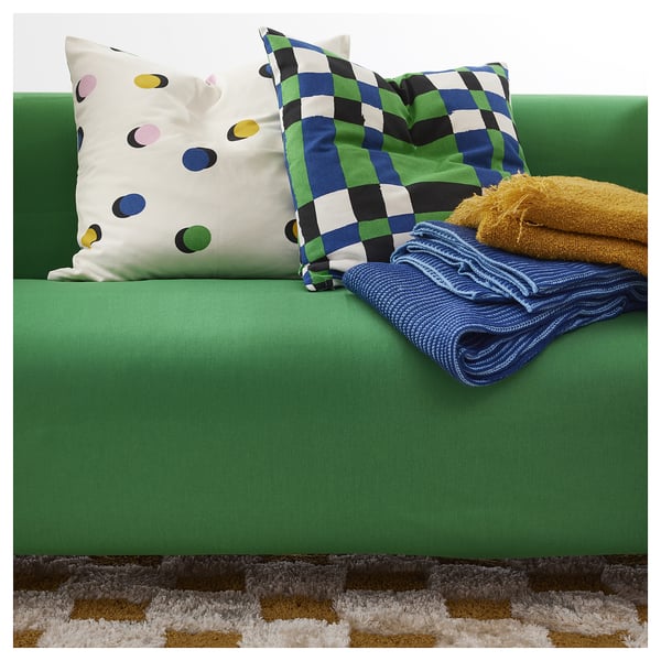 Sofá verde brillante con almohadas punteadas y a cuadros, mantas azules y amarillas, alfombra peluda.
