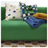 Sofá verde brillante con almohadas punteadas y a cuadros, mantas azules y amarillas, alfombra peluda.