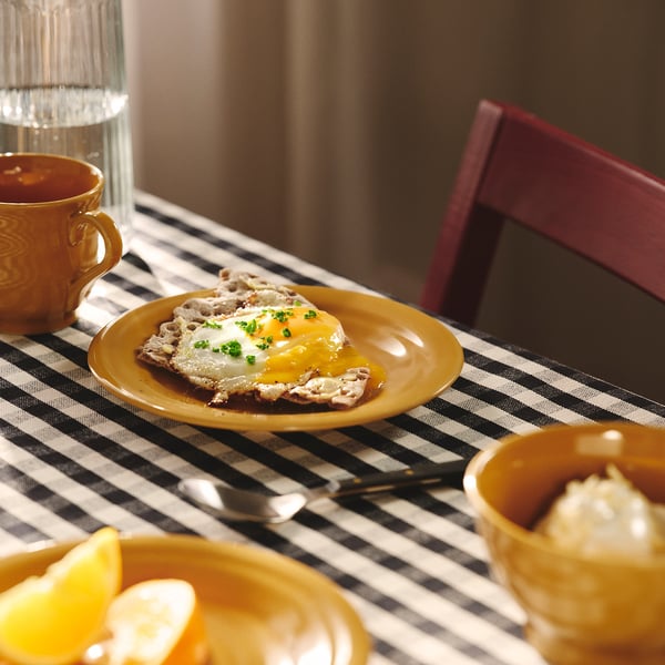 Plato amarillo con huevo frito y hierbas. Rodeado de tazas y vasos sobre un mantel a cuadros.