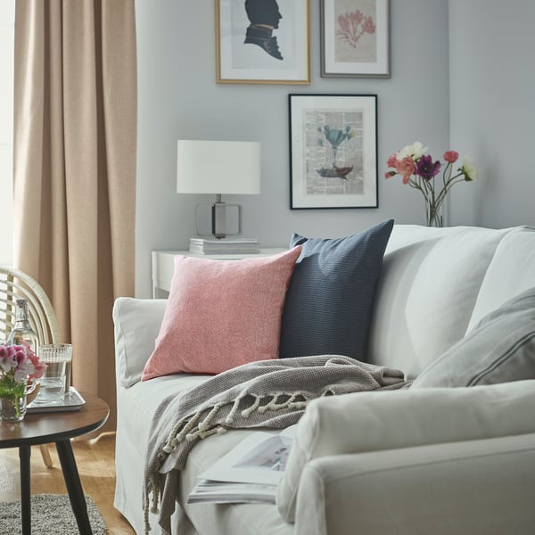 Sala de estar luminosa con sofá blanco, almohadas rojas y azules, mesa auxiliar de madera, lámpara, flores y cortinas grises.