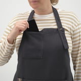 Persona que viste una camisa a rayas y un delantal negro. El delantal tiene cuello ajustable, prácticos bolsillos y lazo para colgar herramientas.