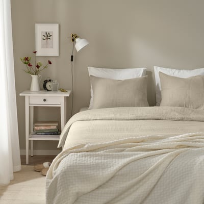 Dormitorio neutro con sábanas blancas y edredón SCHERSMIN. Mesa auxiliar con flores y libros.