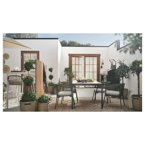 Moderno juego de comedor al aire libre, fondo de la casa blanca. Sillas de metal negro, mesa rectangular de vidrio, plantas en macetas, decoración de patio.