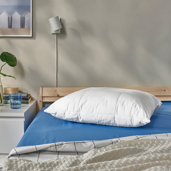 Almohada SENAPSMOTT sobre cama azul, habitación minimalista con planta.
