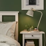 Lámpara SJÖMAKT gris claro en mesita de noche, iluminando la pared verde. La mesa tiene libros, bandeja y decoración. Cama con almohadas a rayas cerca.