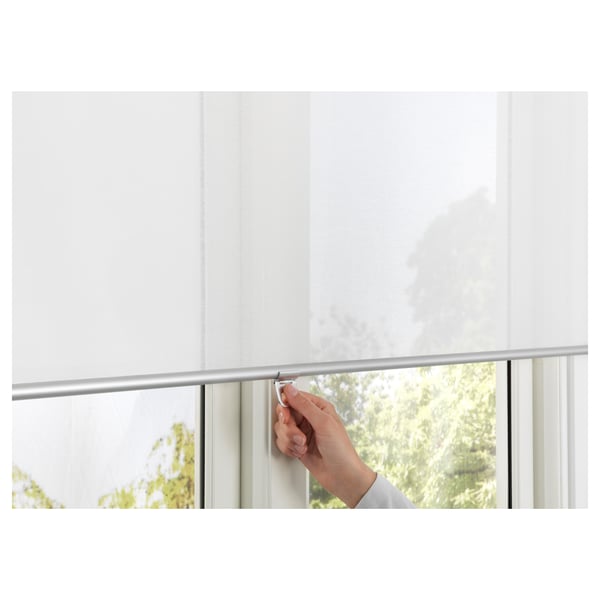 Una mano ajusta una persiana enrollable inalámbrica de cierre suave que filtra la luz a través de una ventana. Seguro para los niños, ofrece privacidad con luz diurna difusa.