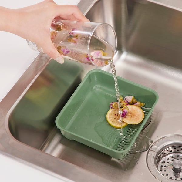 La mano vierte agua del frasco en el colador verde del fregadero con rodajas de limón y flores, lo que demuestra la recolección de desechos de alimentos.