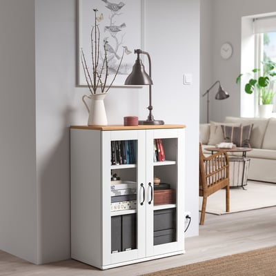 El gabinete de vidrio SKRUVBY en la sala de estar, cerca de la pared gris, sostiene libros, lámparas y decoración.
