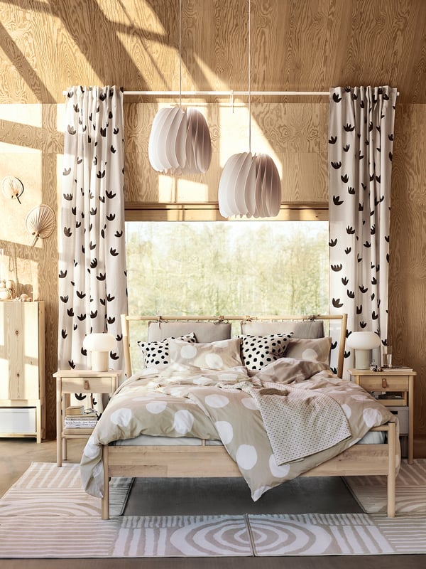 Acogedor dormitorio con decoración de madera clara, lámparas blancas SKYMNINGEN, ropa de cama de lunares, cortinas con coronas, luz natural a través de un gran ventanal.