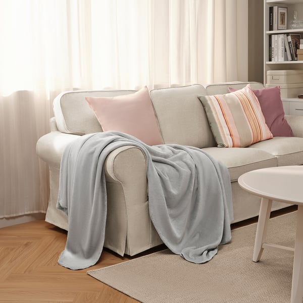 Manta SLÅNGRÅMAL gris sobre sofá con almohadas. Poliéster reciclado, suave, cerca de la ventana con cortinas transparentes.