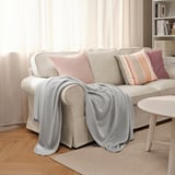 Manta SLÅNGRÅMAL gris sobre sofá con almohadas. Poliéster reciclado, suave, cerca de la ventana con cortinas transparentes.
