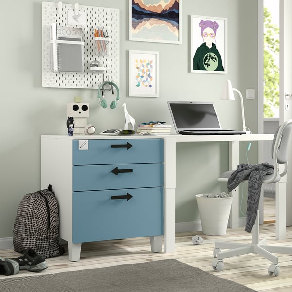 Moderna oficina en casa con escritorio blanco y azul SMÅSTAD, ordenador portátil, silla gris, obras de arte coloridas, auriculares y mochila.