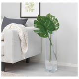 Un jarrón alto y transparente sostiene una sola hoja artificial verde junto a un sofá gris adornado con una almohada y una manta blancas y azul marino.